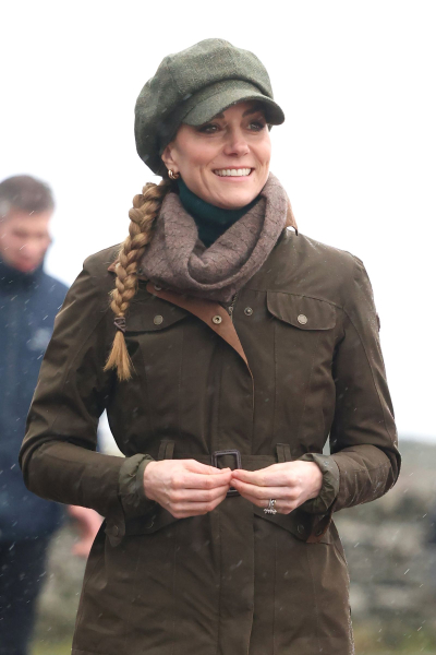 Kate Middleton wears a green jacket and newsboy cap with a scarf. Her hair is styled in a braid.
